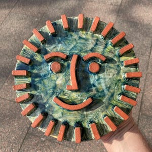 May include: A ceramic sun face plate with a smiling expression. The plate has a blue and green background with an orange sun face. The sun's rays are represented by rectangular orange pieces around the edge. Diameter approximately 25 cm.