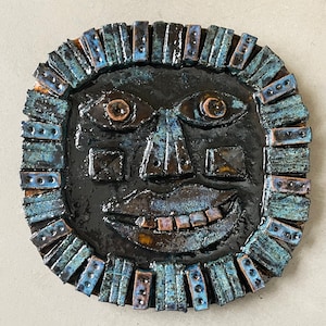 May include: A ceramic sun face wall hanging with a smiling expression. The face is dark with blue and brown accents, including the eyes, nose, and mouth. The sun's rays are made of rectangular blue and brown tiles. A unique piece of art.