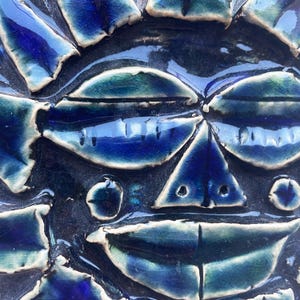 May include: A blue and white ceramic wall hanging featuring a stylised face. The face has a triangular nose, two eyes, and a wide mouth.