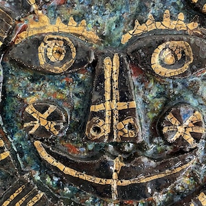 May include: A ceramic wall hanging featuring a stylised face. The face is painted in shades of brown, green, and gold. The eyes are large and round, and the mouth is a wide, open smile. The face is surrounded by a decorative border.