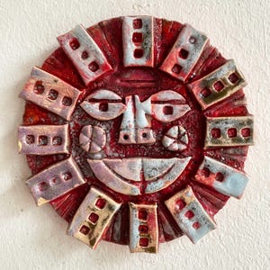 May include: A ceramic sun face wall hanging. The red background features a smiling face with light blue and white accents. The sun's rays are rectangular with a brick-like design.