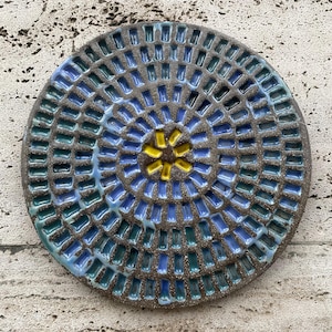 May include: A circular ceramic plate with a mosaic pattern. The plate has a dark brown base, with blue, green, and yellow rectangular tiles arranged in concentric circles. The centre has a yellow starburst design.