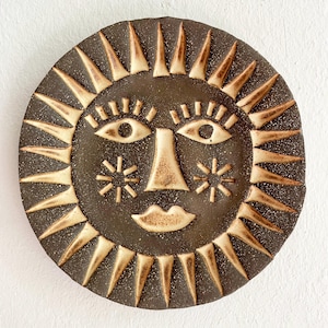 Könnte beinhalten: Eine kreisförmige, dunkelbraune Keramik-Wandplatte mit einem stilisierten Sonnengesicht. Das Design umfasst erhabene, cremefarbene Strahlen am Rand, mandelförmige Augen mit Wimpern, eine dreieckige Nase, sternförmige Wangen und einen kleinen Mund.