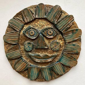 May include: A ceramic sun face wall hanging with a gold and green glaze. The sun has a smiling face with large eyes, a triangular nose, and a wide mouth.