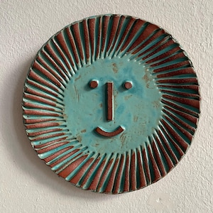 May include: A circular ceramic plate featuring a sun design with a turquoise face and a smiling expression. The sun's rays are reddish-brown, emanating from the face. This decorative plate is a unique piece.