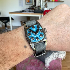 Può includere: Un orologio da polso unico con una cassa quadrata color bronzo e un quadrante blu. L'orologio presenta una lancetta nera e indicatori quadrati. Ha un cinturino in tessuto grigio con chiusura argentata.
