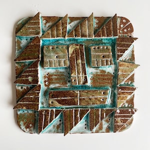 May include: A square ceramic wall art piece with a textured surface. The artwork features a geometric design with brown, turquoise, and white colours. The piece has a unique, handcrafted appearance, with triangular and rectangular shapes.