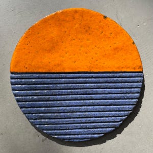 Könnte beinhalten: Eine kreisförmige Wandplatte aus Keramik mit zweifarbigem Design. Die obere Hälfte ist mit einer glatten, glänzenden orangefarbenen Glasur versehen, während die untere Hälfte eine texturierte, matte blaue Glasur mit horizontalen Rillen aufweist.