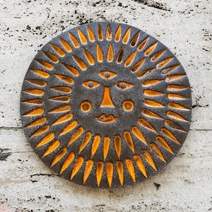Op de afbeelding: Een ronde keramische wandplaat met een gestileerd zonnegezicht en een derde oog op het voorhoofd. Het object is afgewerkt met een donkergrijze matte glazuurlaag, waarbij de zonnestralen, ogen en lippen zijn ingevuld met fel oranje glazuur.