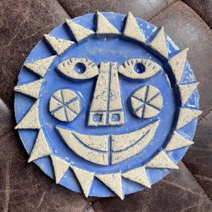 Puede incluir: Una placa de pared circular de cerámica con un rostro de sol estilizado en esmalte azul brillante. El sol presenta un diseño en relieve texturizado de color claro que incluye rayos triangulares, ojos almendrados y una amplia sonrisa.