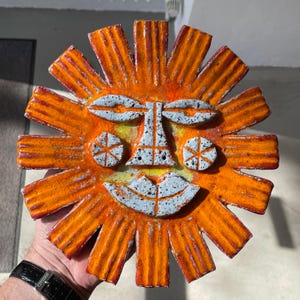 May include: A ceramic sun face sculpture in orange and yellow. The sun has a smiling face with white and blue details. The sun's rays radiate outwards, forming a bright design. The sculpture is a decorative item.