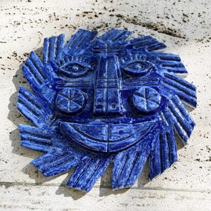 Könnte beinhalten: Eine kreisförmige blaue Wandplatte aus Keramik mit einem stilisierten Sonnengesicht, das einen breiten lächelnden Mund, zwei kreisförmige Wangendetails und strahlenförmige Sonnenstrahlen um den gesamten Rand aufweist.