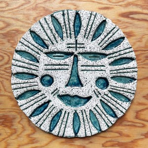 May include: A circular ceramic sun face with a speckled white surface and teal blue detailing. The sun features a smiling mouth, a long nose, and oval eyes. The sun's rays are depicted by lines and leaf-shaped patterns.