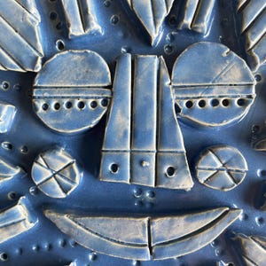 May include: Close-up of a blue ceramic art piece with a face-like design. The artwork features geometric shapes, including circles, rectangles, and triangles, with a textured surface and small circular indentations. The piece is a vibrant shade of blue.