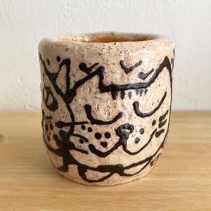Può includere: Una tazza in ceramica beige con superficie strutturata e segni astratti neri. L'interno della tazza è arancione. La tazza ha una forma cilindrica e sembra fatta a mano.