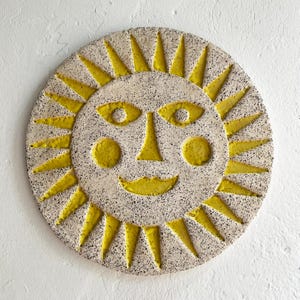May include: A circular ceramic sun wall decoration with a smiling face and yellow rays. The sun's face is off-white with black speckles, and the facial features and rays are bright yellow. A decorative piece for the home.