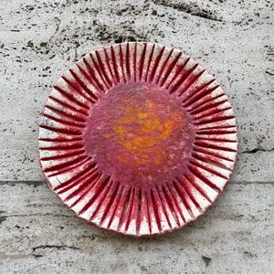 May include: A handcrafted ceramic plate with a distinctive design. The plate has a central area with orange and yellow tones, encircled by a textured, reddish-pink surface. The outer rim features red, vertical lines against a white background.