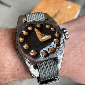May include: A handcrafted wristwatch featuring a dark watch face with gold hands and hour markers. The watch has a grey fabric strap and a metallic case. The watch face is black with gold accents. The watch is worn on a wrist.