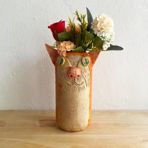 May include: A handcrafted ceramic vase in the shape of a cat's face, with orange and beige hues. The vase has large, cartoonish eyes, a pink nose, and a smiling mouth. It is filled with a colourful arrangement of artificial flowers, including a red rose.