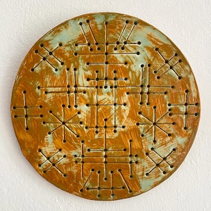 Peut inclure: Une pièce décorative ronde en céramique à surface texturée. Elle présente un motif géométrique de lignes et de points. Les couleurs dominantes sont l'orange et le bleu sarcelle, avec un aspect rustique et vieilli. Environ 25 cm de diamètre.