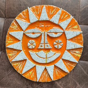 May include: A ceramic sun face with an orange centre and white triangular rays. The sun has a smiling face with detailed features, including eyes, a nose, and a mouth. The sun is a decorative item.