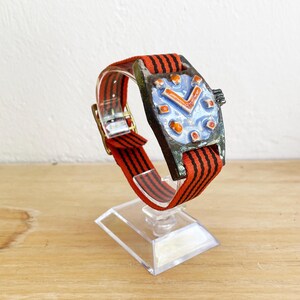 May include: A distinctive wristwatch featuring a rectangular, dark-toned face and a blue dial with orange detailing. The watch is paired with a red and black striped strap. It is presented on a clear stand.