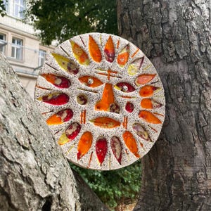 May include: A circular ceramic sun face sculpture with a speckled white background. The sun's features are made from orange, red, and yellow leaf-shaped pieces. The sculpture is mounted on a tree trunk.