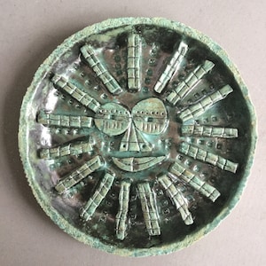 May include: A green ceramic plate featuring a raised design of a sun face. The sun has a smiling mouth, eyes, and a nose. The sun's rays are made of small squares.