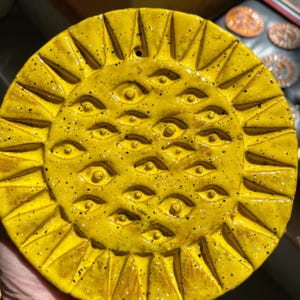 Puede incluir: Una placa de pared circular de cerámica amarilla con un diseño similar al sol con rayos triangulares radiantes alrededor del borde. El centro está densamente decorado con múltiples motivos de ojos en relieve, cada uno con una pupila central.