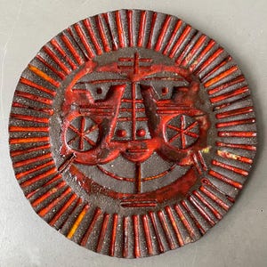 Puede incluir: Un plato decorativo redondo de cerámica con un diseño de cara solar. El plato presenta una base marrón oscuro con una cara roja vibrante y líneas radiales. La cara tiene rasgos geométricos y formas de rueda a ambos lados.