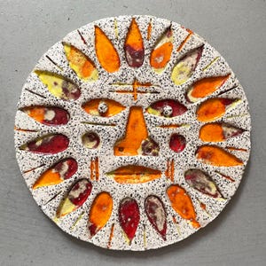 Könnte beinhalten: Eine runde, dekorative Keramikplatte mit einem sonnenähnlichen Gesichtsdesign. Die Platte hat einen weißen, gesprenkelten Hintergrund mit orangefarbenen, gelben und roten Akzenten. Das Gesicht hat Augen- und Munddetails, und die Sonnenstrahlen sind stilisiert.