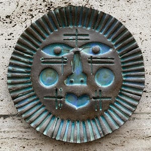 May include: A circular, bronze-coloured wall sculpture with a stylised face design. The face features blue accents for the eyes, nose, cheeks, and mouth. The outer edge has a sunburst pattern.