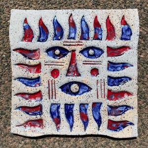 May include: A square ceramic tile with a face design. The face has two blue eyes, a red nose, and red and blue accents. The tile has a textured, off-white background. It is likely a decorative art piece, possibly for wall display.