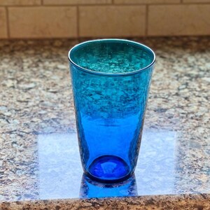 May include: A tall, textured glass in shades of blue, transitioning from a deep azure at the base to a vibrant turquoise at the rim. The glass is empty and sits on a speckled countertop.