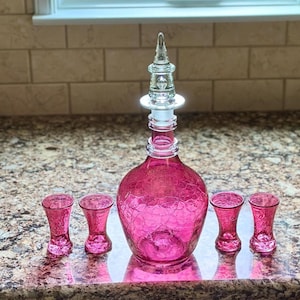 May include: A pink glass decanter with a crackle finish, topped with a clear stopper, and surrounded by four matching shot glasses. The set is displayed on a reflective surface, suggesting a barware or home decor theme.