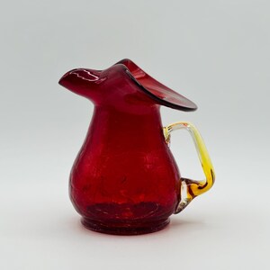 May include: A small, ruby-red glass pitcher with a crackle finish. The handle is yellow and clear, and the spout has a flared, curved design. The pitcher is likely vintage and suitable for serving beverages.