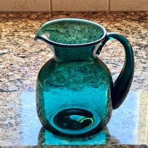 May include: A teal-colored glass pitcher with a rounded body and a curved handle. The pitcher has a spout for easy pouring and is made of transparent glass, allowing light to pass through. The pitcher is likely used for serving beverages.