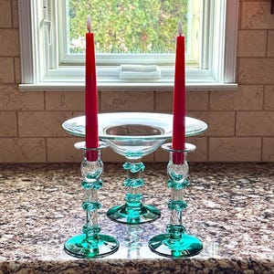 May include: A decorative centerpiece featuring two red candles and two teal glass candle holders. The centerpiece includes a clear glass bowl. The candle holders have a decorative design and are placed on a granite countertop.