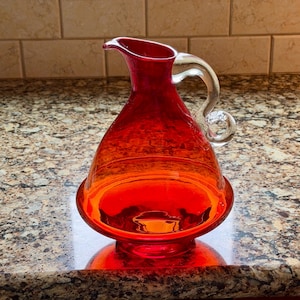 May include: A vibrant red glass pitcher with a clear glass handle. The pitcher has a wide base and a narrow spout. The glass has an orange gradient at the base. The pitcher is likely used for serving beverages.