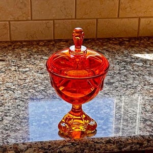 May include: An orange-colored, lidded candy dish. The glass dish has a pedestal base and a decorative handle on the lid. The dish is sitting on a granite countertop.