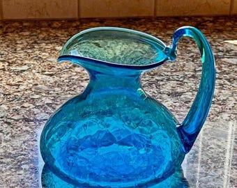May include: A vibrant blue glass pitcher with a distinctive handle and a wide, flared rim. The pitcher has a textured surface and a rounded base. The handle curves gracefully from the rim to the body, creating an elegant silhouette. The pitcher is likely used for serving beverages.