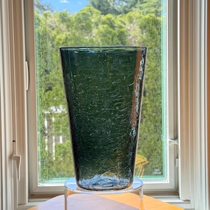May include: A tall, tapered glass vase in a deep forest green hue. The exterior surface features a distinct crackle texture, creating a network of fine, irregular lines across the translucent body. The vase has a wide, open rim and a narrow base.