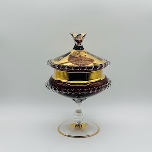 May include: A vintage glass candy dish with a burgundy base and gold accents. The lid features a gold design with a scene of people in a garden.