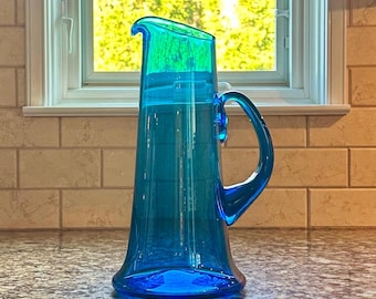 May include: A tall, turquoise-blue glass pitcher with a curved handle. The pitcher has a flared base and a slightly angled spout. The background includes a window with a view of green foliage and a light-colored tiled wall.