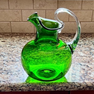 May include: A vibrant green glass pitcher with a clear, curved handle. The pitcher has a round base and a flared rim. The glass has a textured appearance, and the handle is attached at the top and bottom of the pitcher.