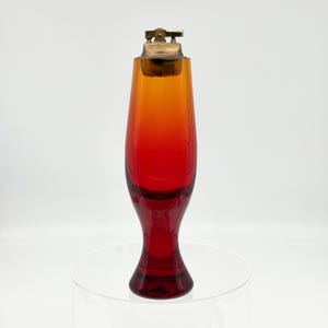 May include: A tall, slender table lighter featuring a vibrant gradient glass body that transitions from a deep red base to a bright orange top. The lighter is topped with a metallic gold-toned ignition mechanism, showcasing a sleek, elongated silhouette.