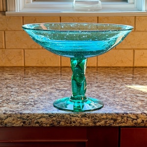 May include: A teal glass pedestal bowl featuring a wide, shallow basin with visible air bubbles throughout the material. The bowl is supported by a thick, twisted glass stem that connects to a circular base, creating a stable, elevated structure.