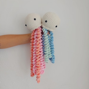 Pode incluir: Dois brinquedos de água-viva de crochê com corpos brancos e tentáculos coloridos. Uma água-viva tem tentáculos rosa e laranja, e a outra tem tentáculos azuis e verdes.