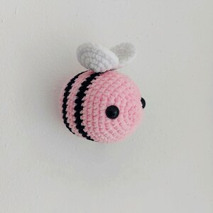 Pode incluir: Uma abelha de crochê rosa com listras pretas e asas brancas. A abelha tem olhos pretos de botão.