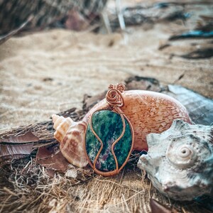 May include: A green and pink gemstone pendant wrapped in copper wire. The pendant is sitting on a bed of brown and white shells.